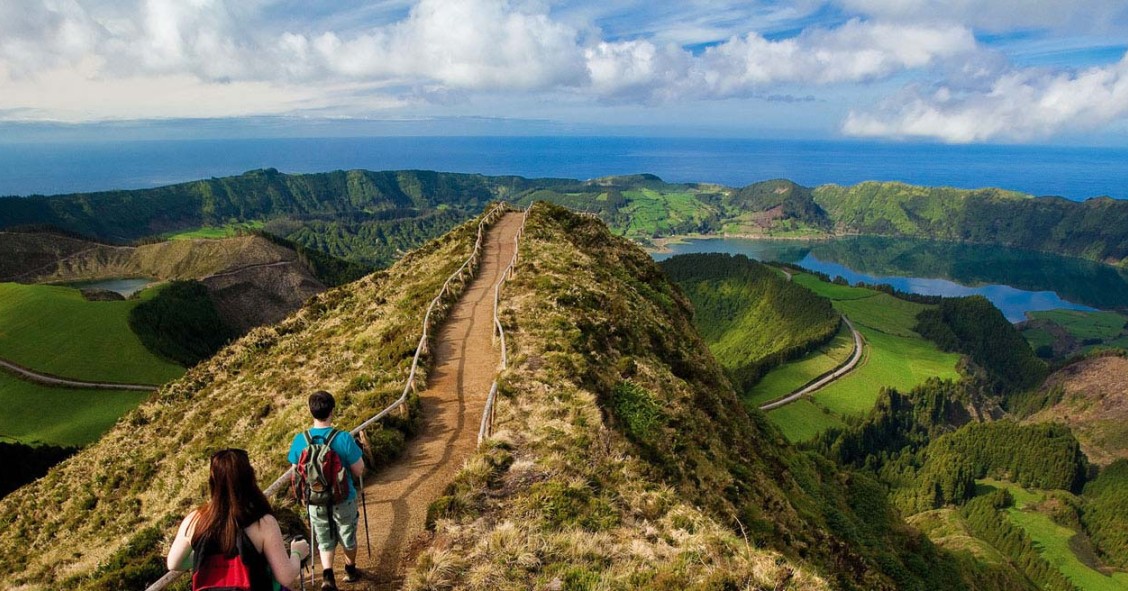 Imagem de pessoas a caminhar num trilho junto a uma lagoa, num cenário de pura natureza verde.