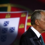 Imagem de Marcelo Rebelo de Sousa de perfil, com bandeira de Portugal como pano de fundo.